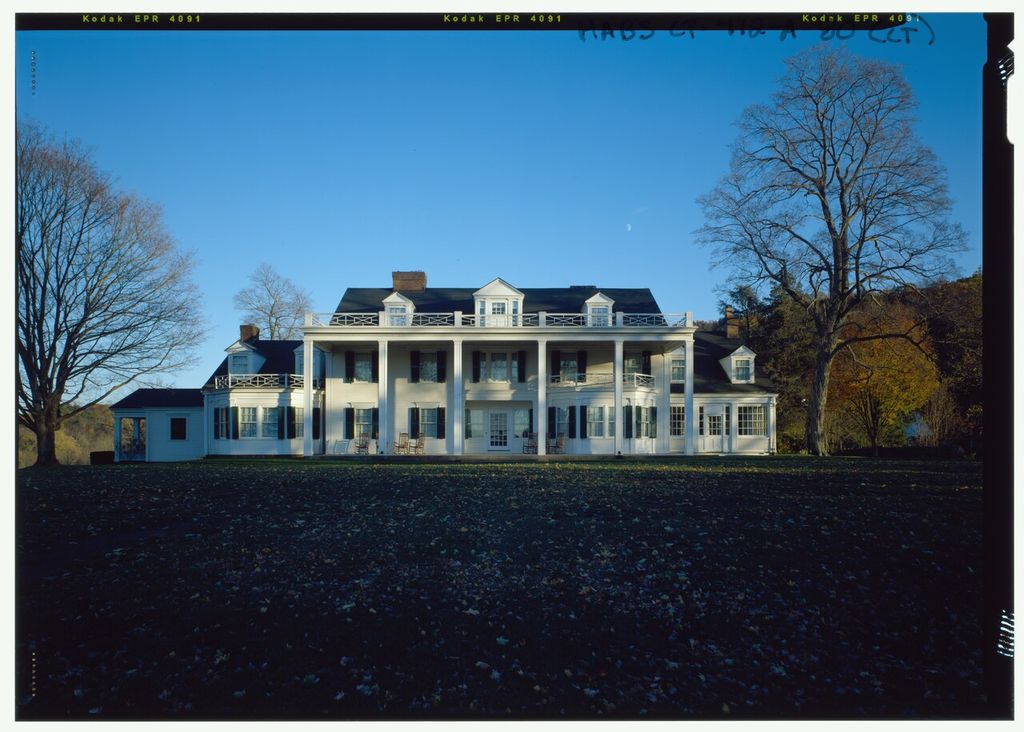 A white house has a long two-storey colonnade, roof dormers, balustrades and lower side wings beyond a broad lawn.