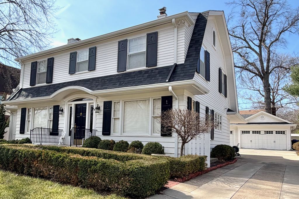 A white house has a broad upper dormer, a steep gambrel end, black shutters and a curved canopy over its central entrance, beside a driveway leading to a garage.