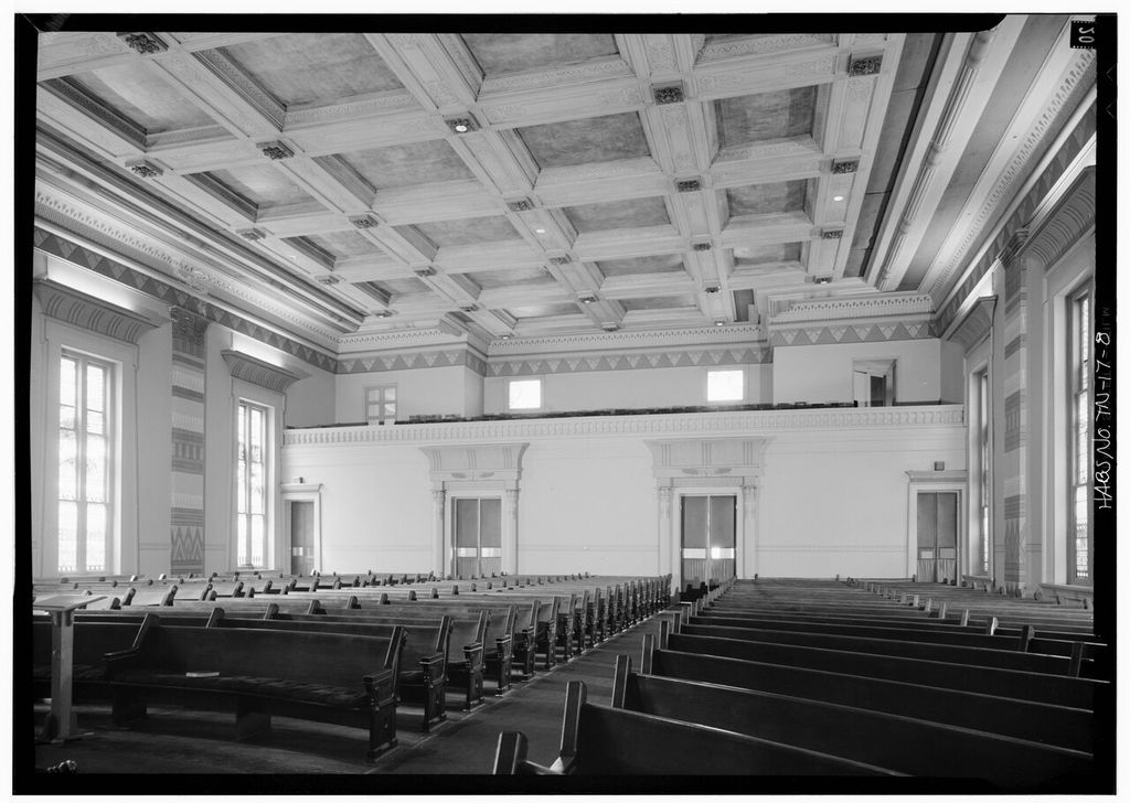 Pews face away from a rear gallery beneath a coffered ceiling, with patterned pilasters and decorated door surrounds.