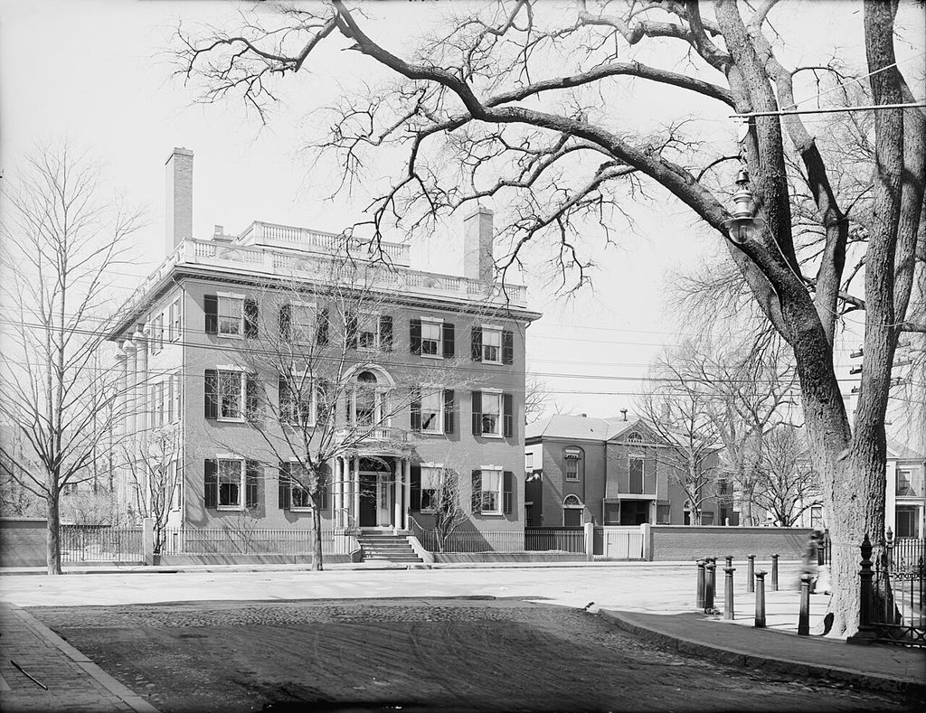 A three-storey house with a central entrance and roof balustrade faces a tree-lined street.