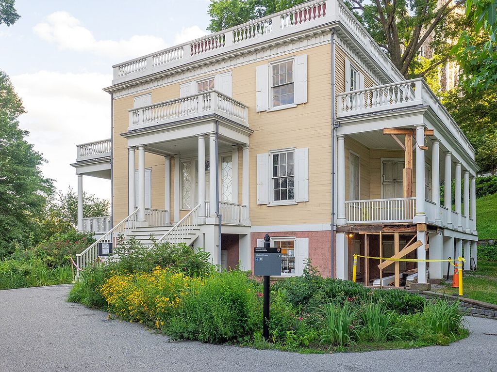 A pale clapboard house has a columned central porch and balanced windows, with temporary supports at one side.