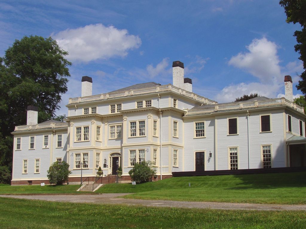 A pale clapboard country house has projecting window bays, a central doorway, lower side ranges and roof balustrades.