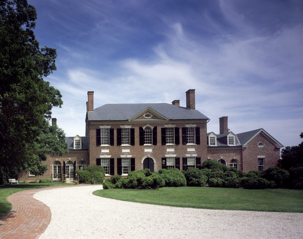 A brick country house with a central arched entrance is joined to lower ranges beyond a curving drive.
