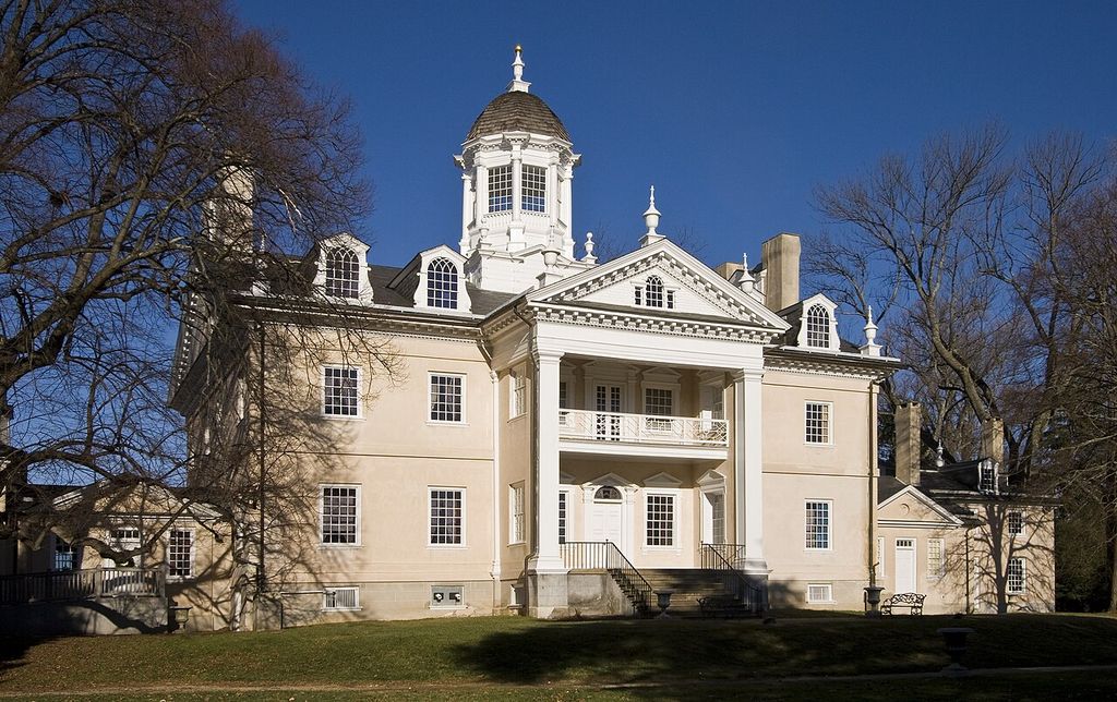 A pale house with a central portico, pediment and cupola, with parts of lower wings visible behind bare trees.