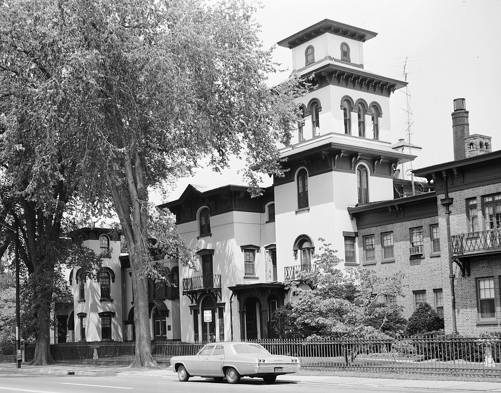 A tall square tower with arched windows and broad bracketed eaves rises above irregular house wings beside a street.