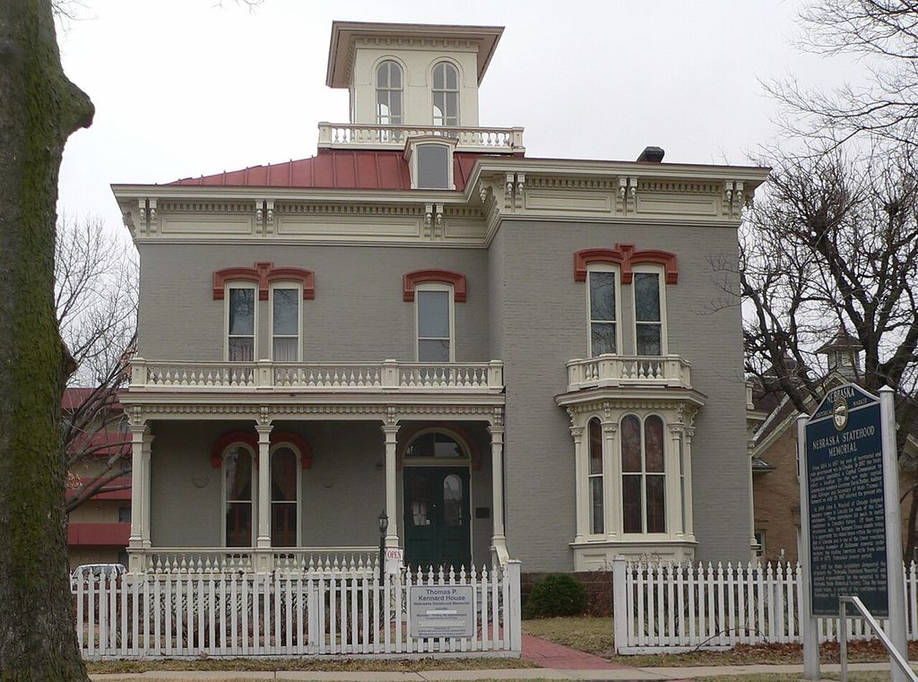 A cupola rises above broad bracketed eaves, tall hooded windows and an ornamental porch behind a picket fence.