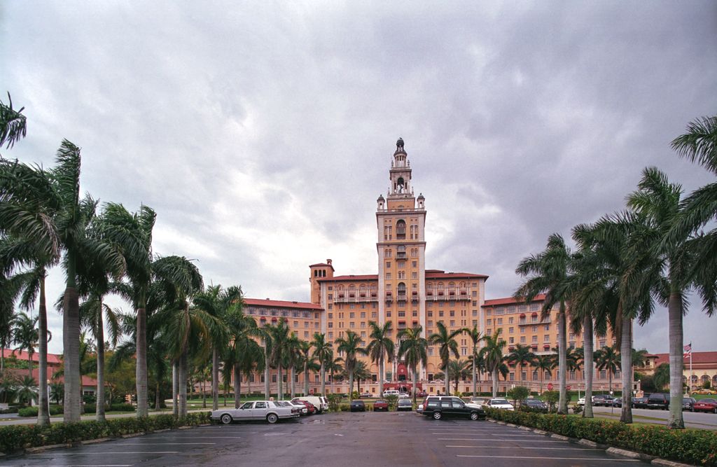 A tall ornamental hotel tower rises between broad wings beyond palms and a parking area.