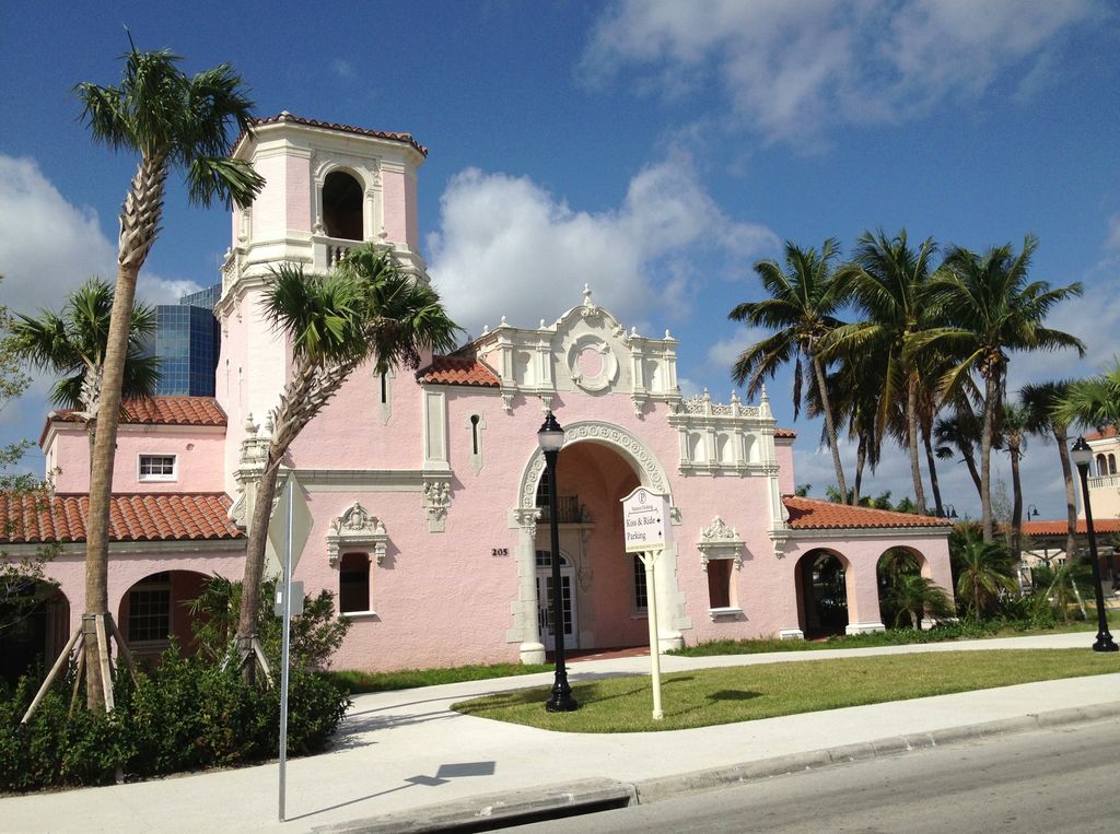 A pink station has an ornate arched entrance, a corner tower, tiled roofs and shaded arcades beside palms.