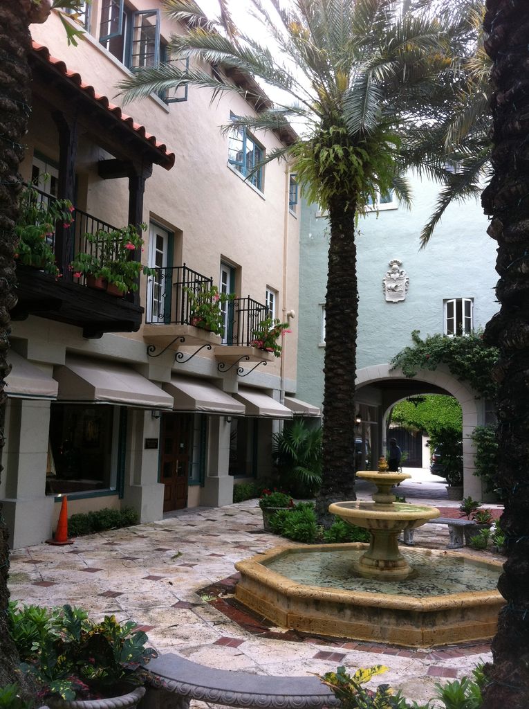Shop fronts and balconies overlook a paved court with a fountain and palms, leading through an arch to another passage.