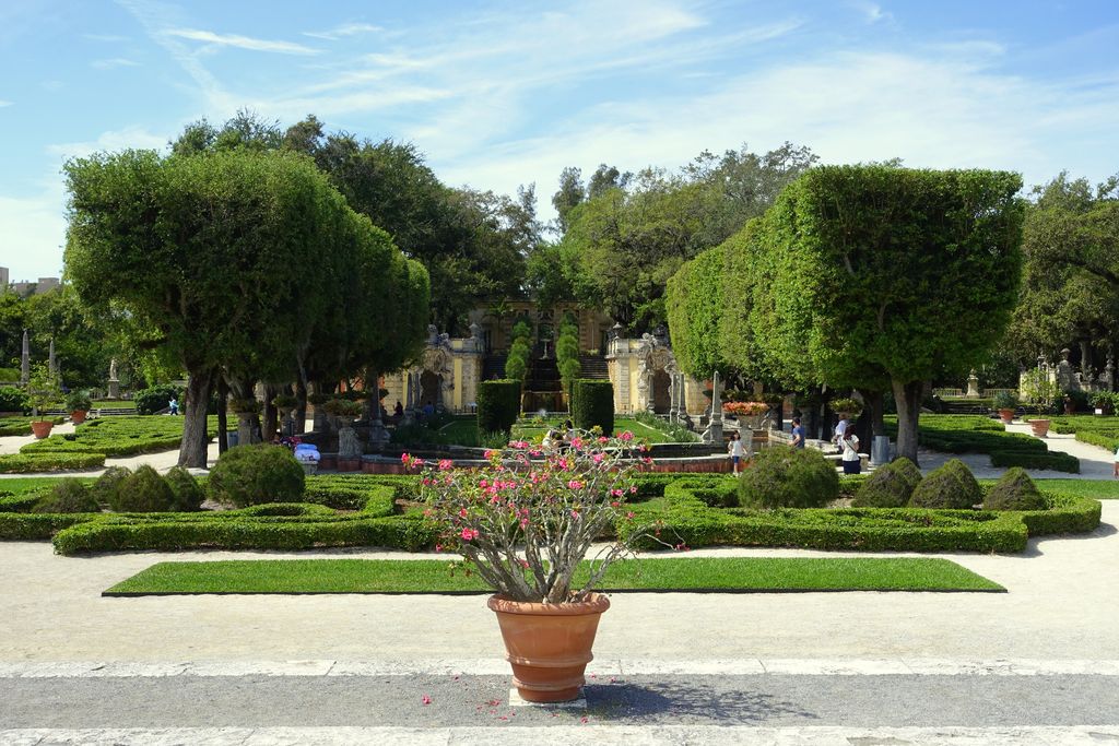 Geometric hedges and paths form an axis between clipped trees toward distant garden steps and stone structures.