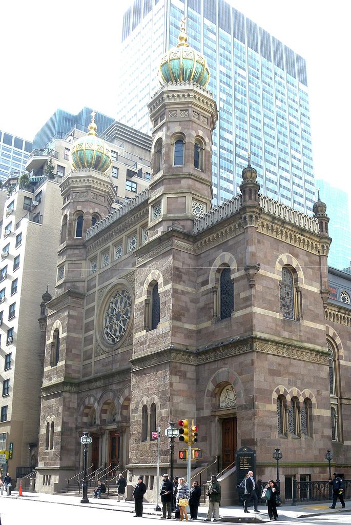 Twin domed towers and banded masonry frame a circular window and arched entrances among taller city buildings.