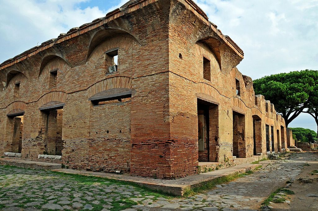 Corner of a brick apartment building at Ostia with upper windows and broad lower openings beside a paved street.