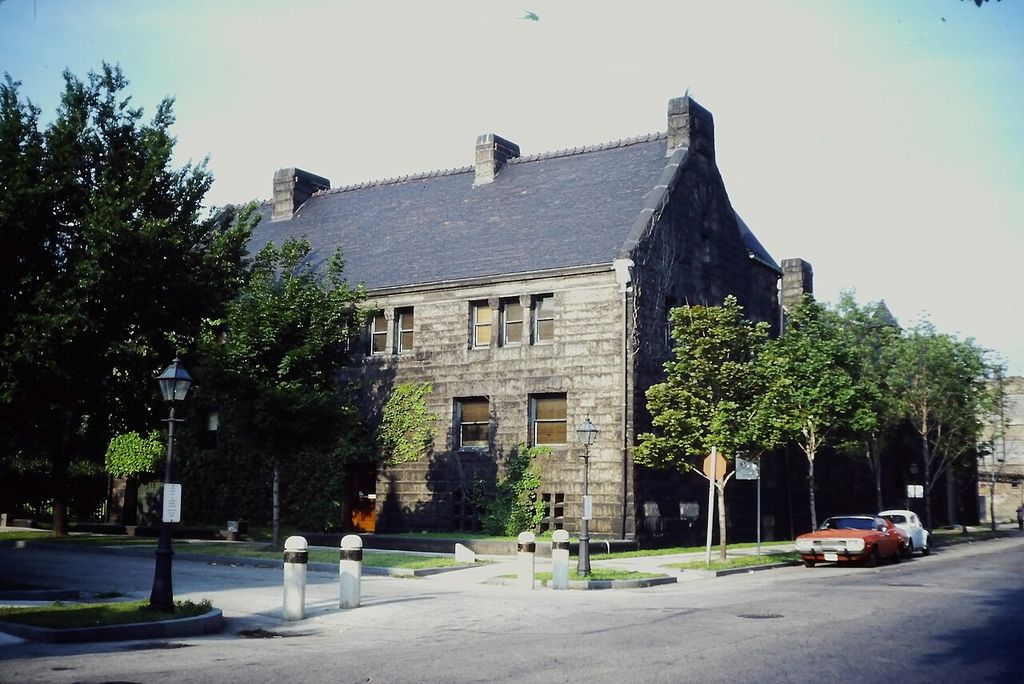 A steep-roofed stone house presents a broad wall with relatively few openings toward a street corner.