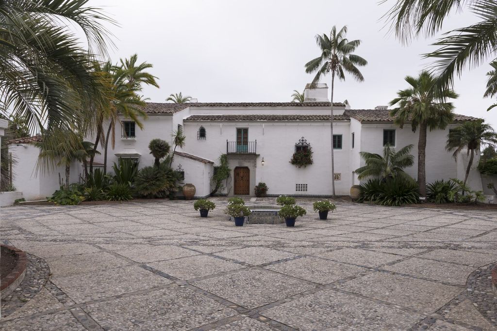A paved court and small fountain lead to a white house with tiled roofs, a balcony and ironwork among palms.