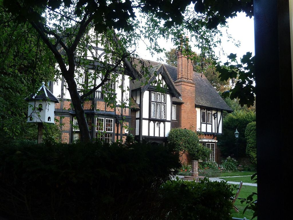 A partially screened house has timbered projecting bays, patterned brick infill, steep gables and a tall chimney.