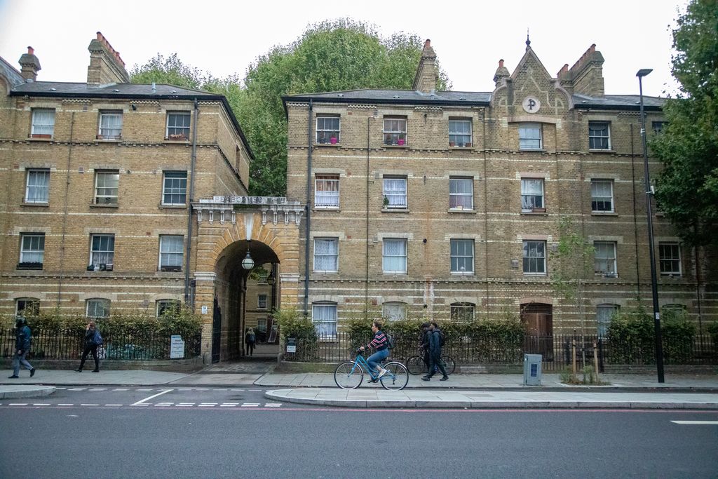 Four-storey yellow-brick housing blocks with repeated sash windows flank a stone archway, with pedestrians and a cyclist in front.