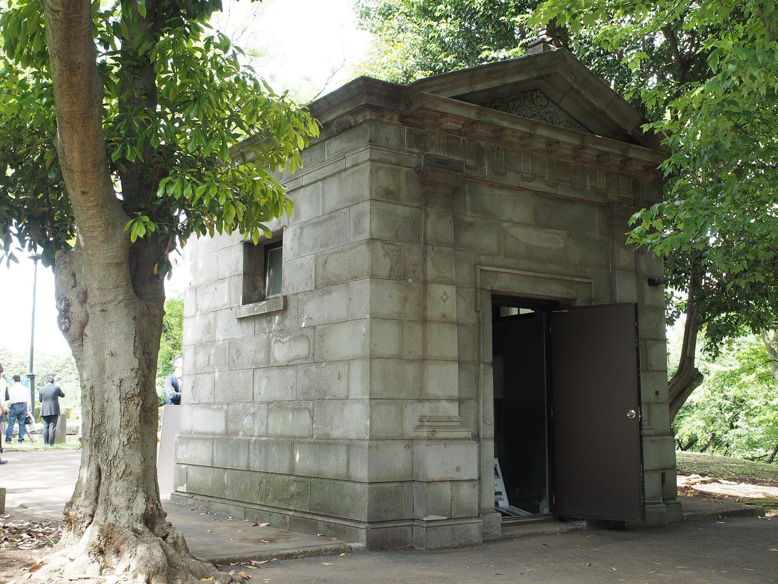 Small stone classical-style housing for the Japanese Vertical Datum among mature trees, with its door standing open.