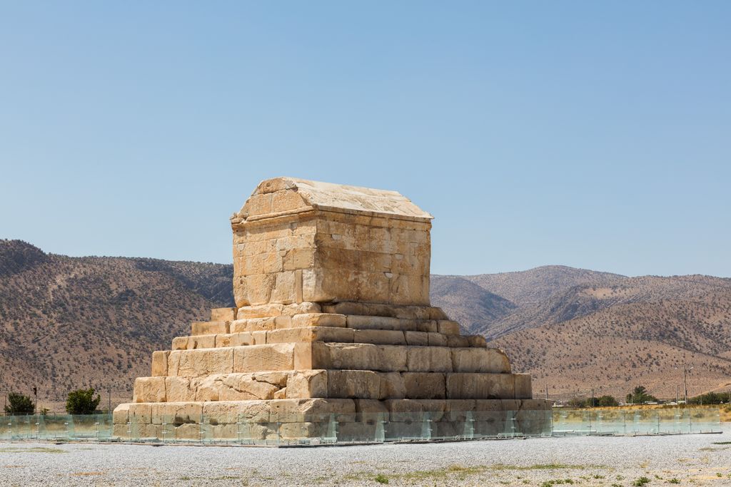 A small gabled stone chamber stands on six broad steps in an open gravel plain before low brown mountains.