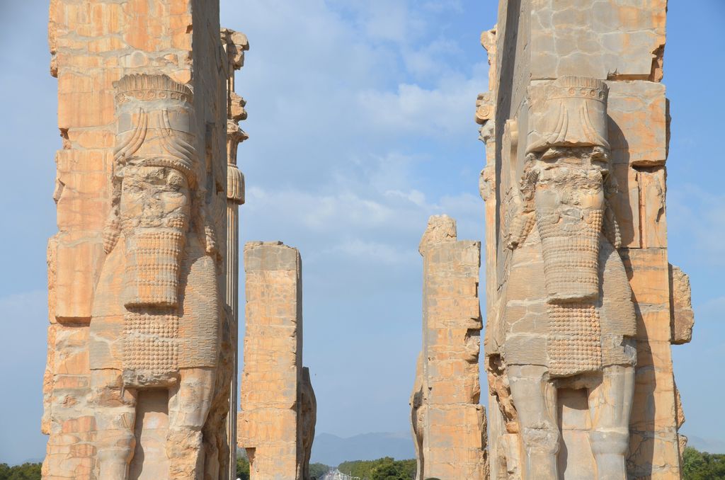 Two tall weathered stone piers carved with winged human-headed bulls frame blue sky and ruined columns beyond.