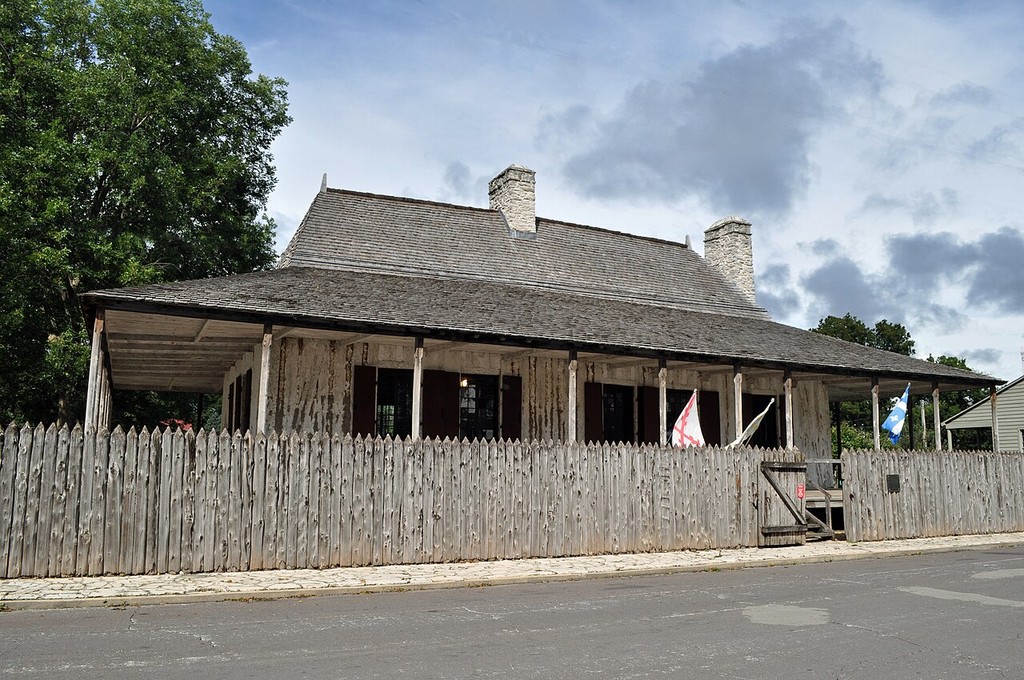 A steep hipped roof extends over a long gallery beside vertical timber wall members and a palisade.