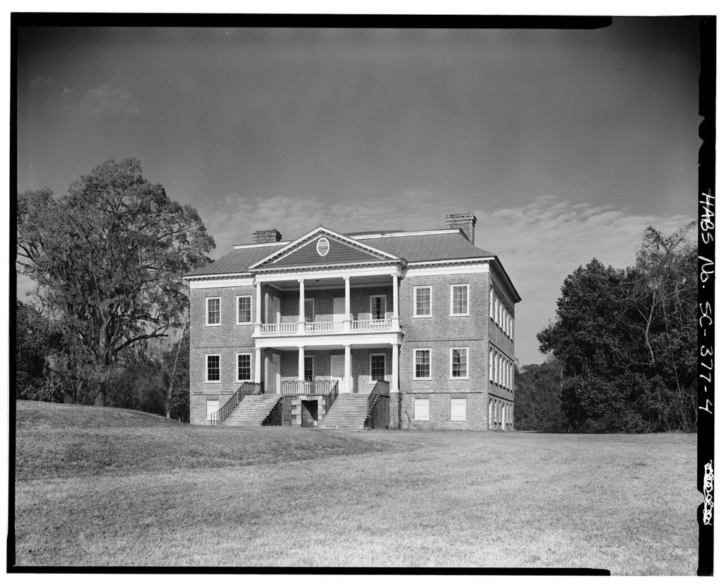 A brick house has a two-storey columned portico, triangular pediment, paired entrance stairs and raised basement.