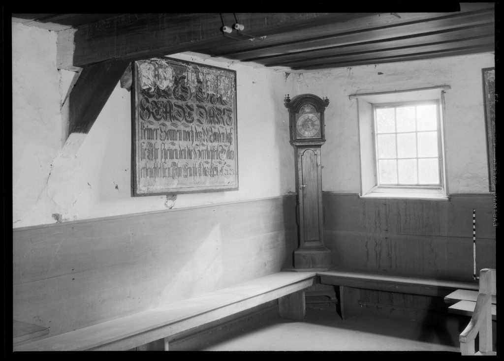 A monochrome interior shows a timber ceiling and brace, wall benches, a window, clock and inscribed panel.