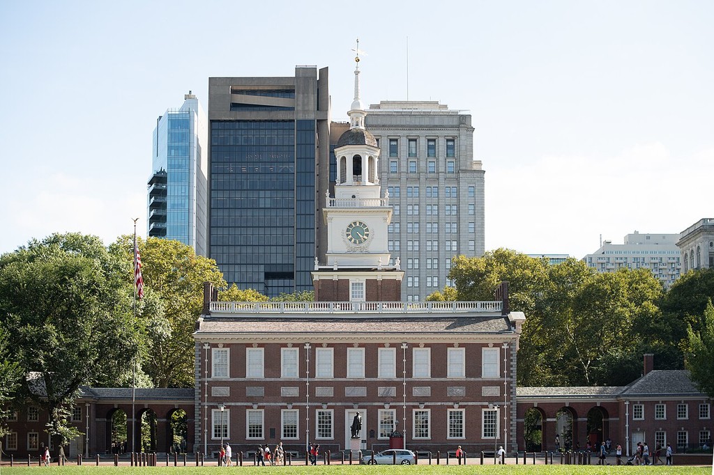A brick institutional front and connecting arcades stand below a clock tower and pale steeple, with later city buildings behind.