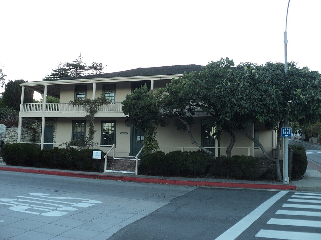 A long upper balcony with slender posts runs above the ground-floor rooms of a two-storey building, partly hidden by trees.