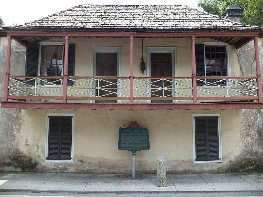A two-storey house has a broad covered timber balcony above a pale plastered ground floor.