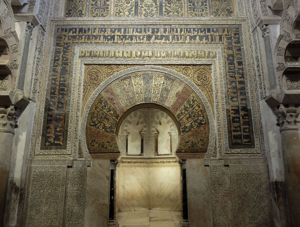 A richly patterned horseshoe arch with gold mosaics and calligraphy frames a small doorway into a pale chamber.