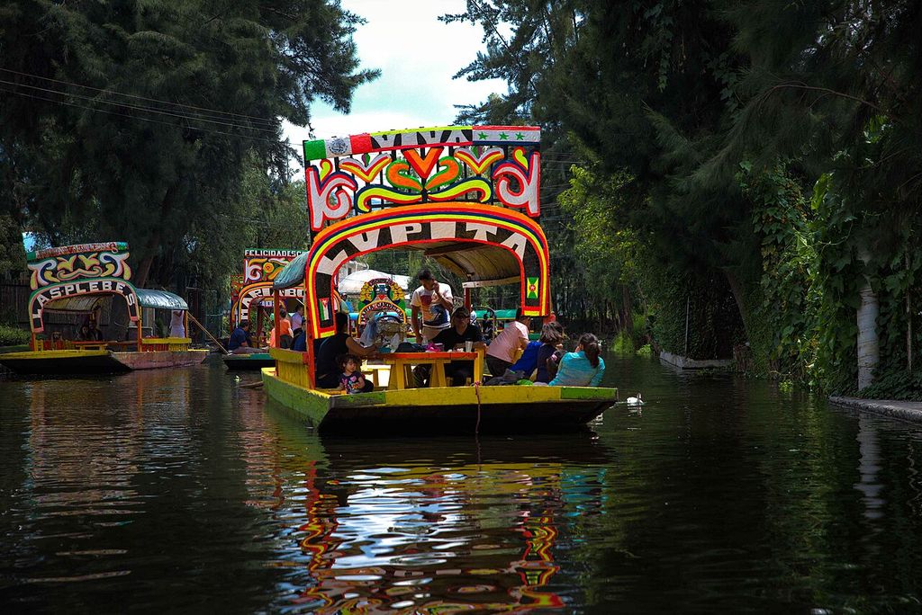Brightly painted passenger boats travel along a narrow shaded canal between dense trees, fences and waterside vegetation.