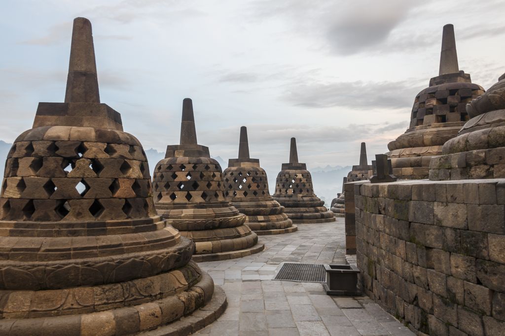 Rows of dark bell-shaped perforated stone stupas stand on stepped terraces beneath an overcast sky.