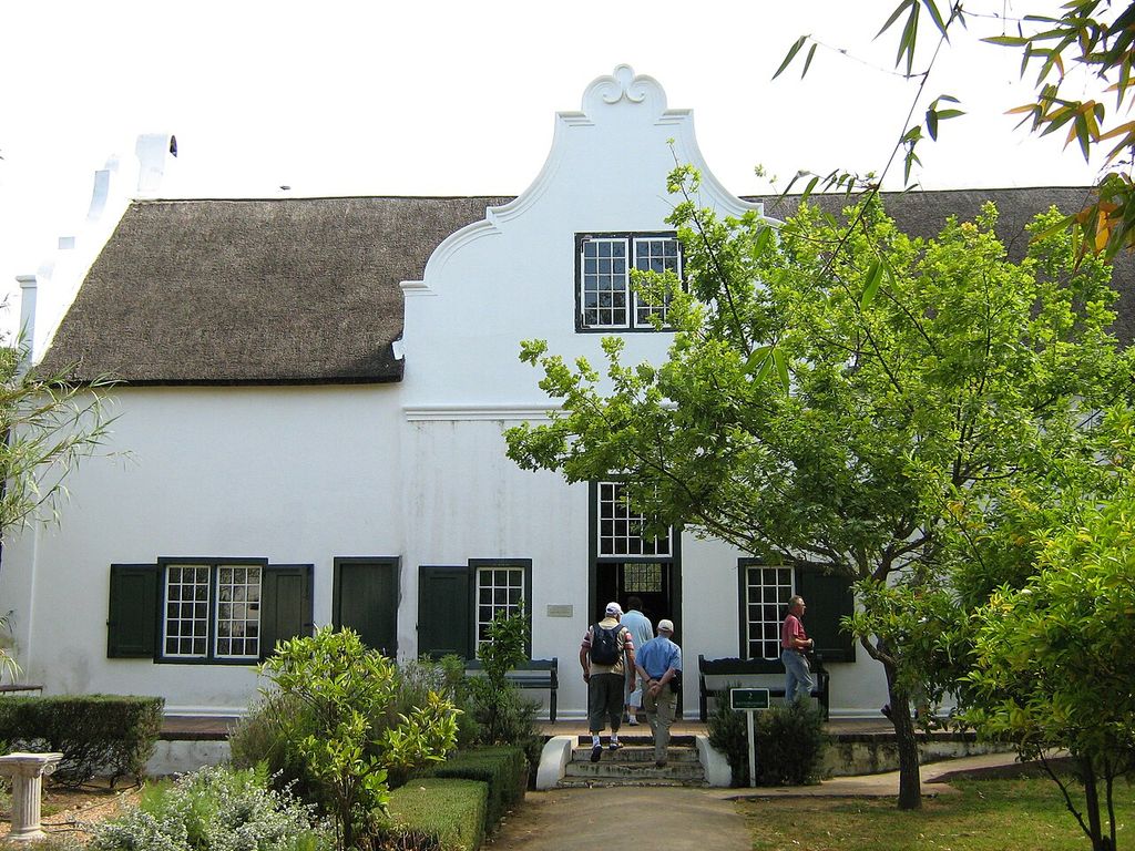 A curved centre gable, thatched roof and shuttered windows face a garden path, with a tree partly covering the right side.