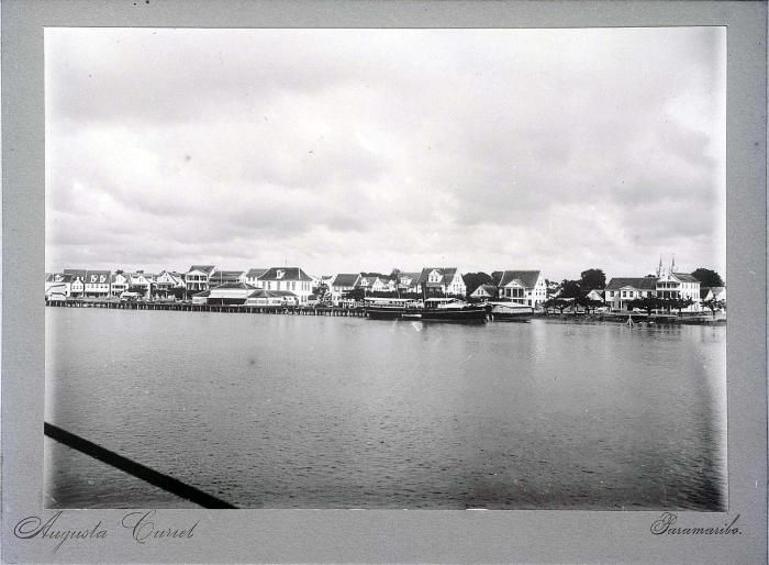 A historic monochrome panorama shows waterfront buildings, palms, a landing and small boats, with the source border and studio mark retained.