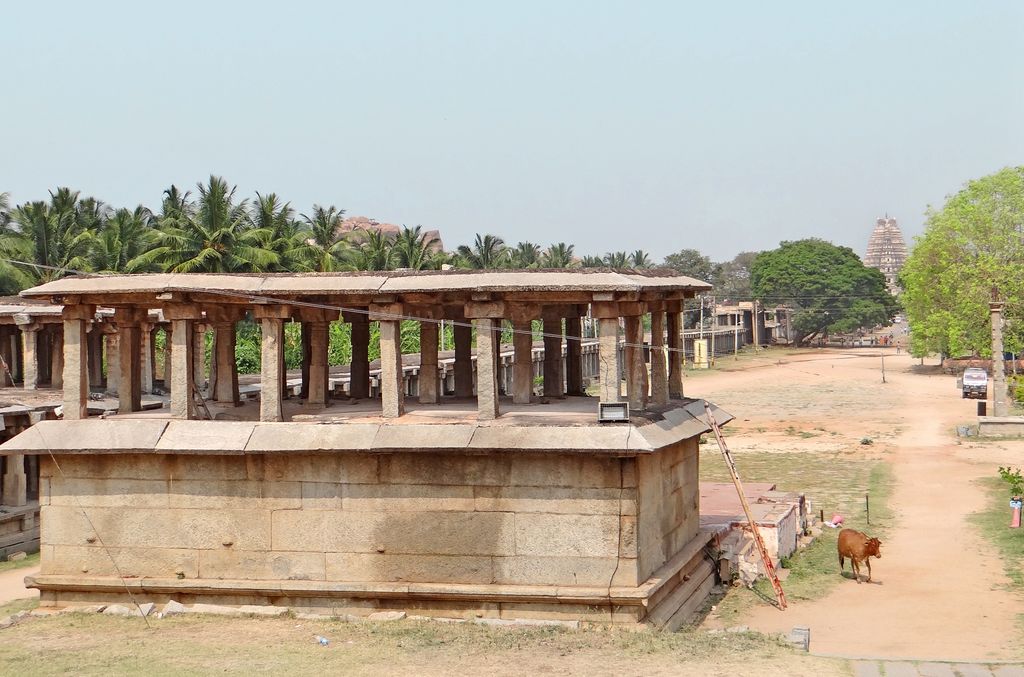 A long stone colonnade borders a broad earthen route lined by palms, with a distant temple tower and scattered animals and vehicles.