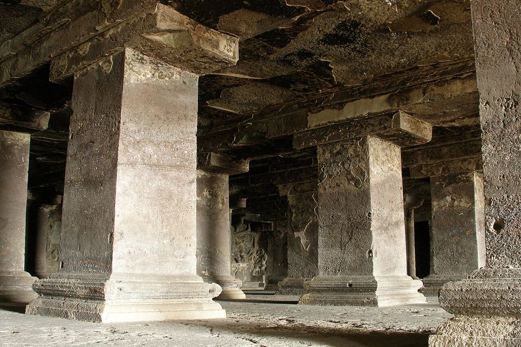 Rows of heavy square basalt columns and carved bases support a dark rock-cut ceiling in a cave hall.