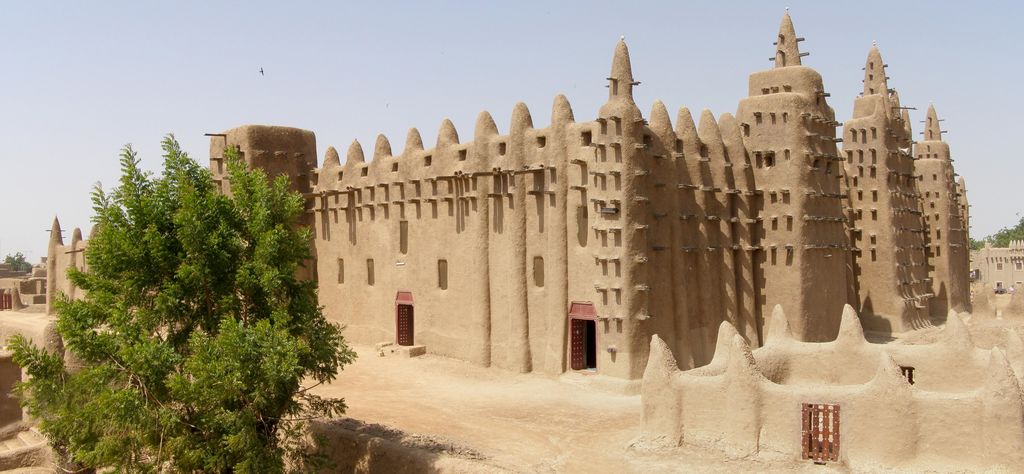 A large ochre earthen mosque with rows of projecting timbers and three tall minaret forms rises behind trees and a low wall.