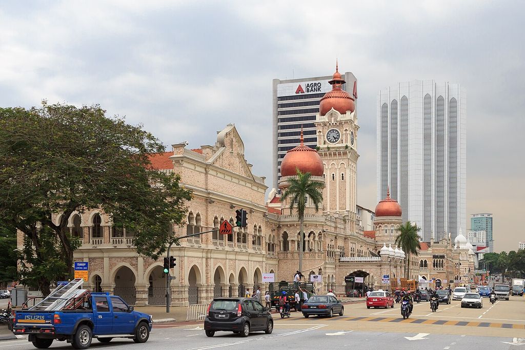 A long arcaded office front with domes and a central clock tower faces a street, with later tall buildings behind.