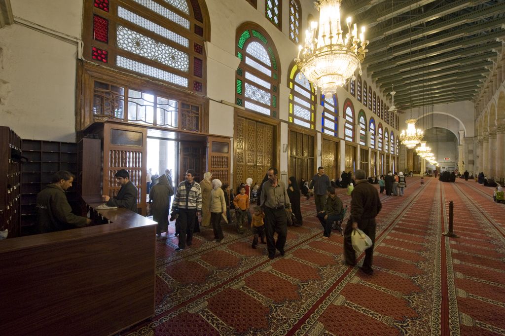 A long carpeted mosque aisle runs beneath repeated dark roof beams, with columns, bright doorways and visitors along the side.