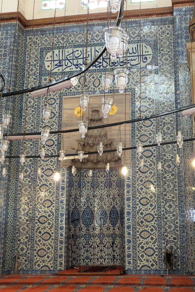 A tall stone mihrab with a carved hood stands within a wall densely covered in blue-and-white floral tiles and inscriptions.