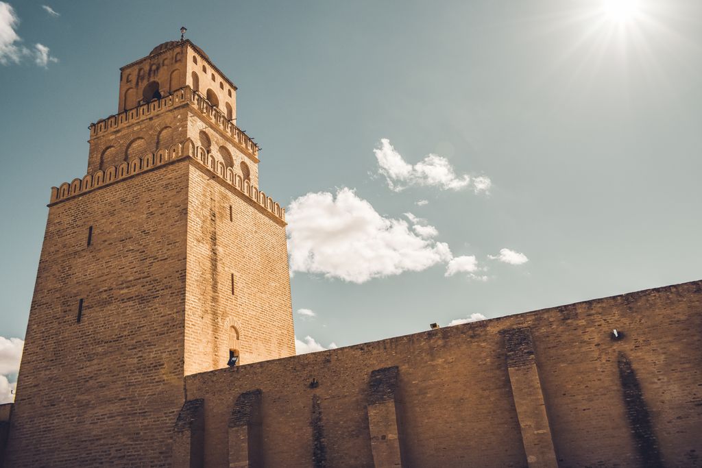 A tall three-stage square stone minaret rises behind a buttressed enclosure wall beneath a dark blue sky.