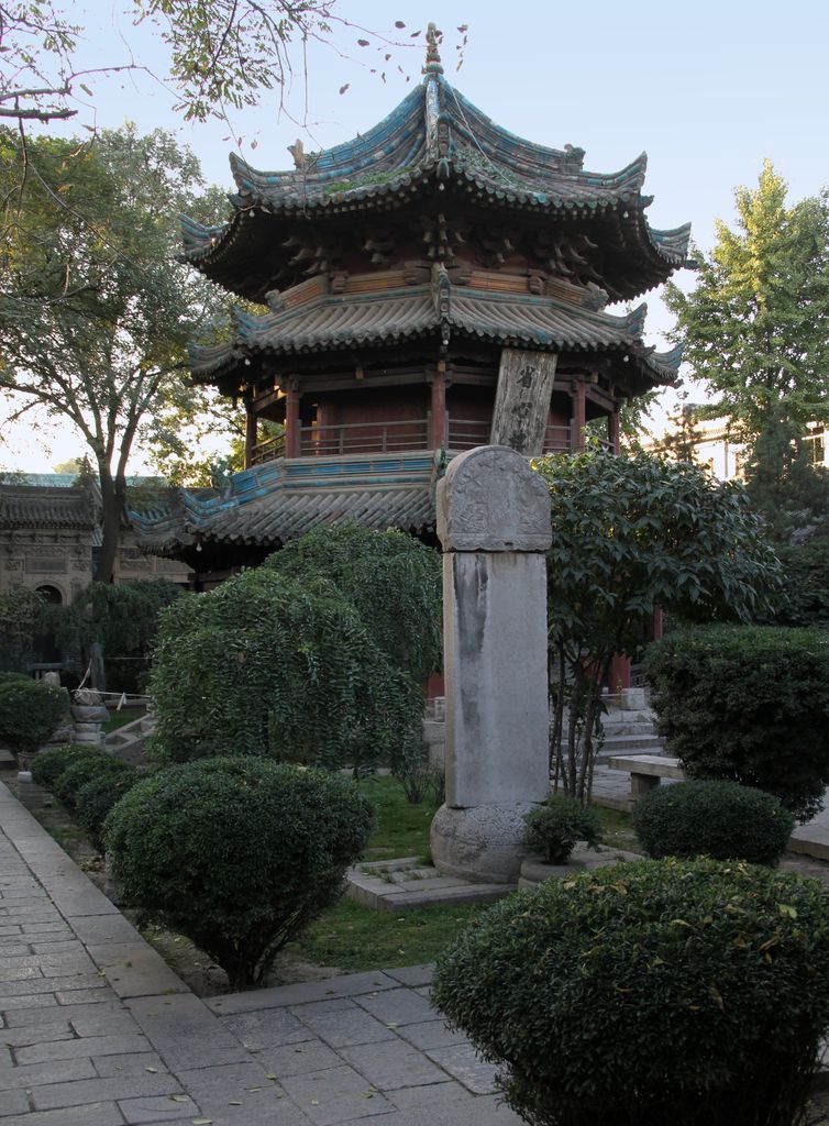 A tall octagonal timber pavilion with several dark tiled roofs and upturned eaves rises among trees behind a stone stele.