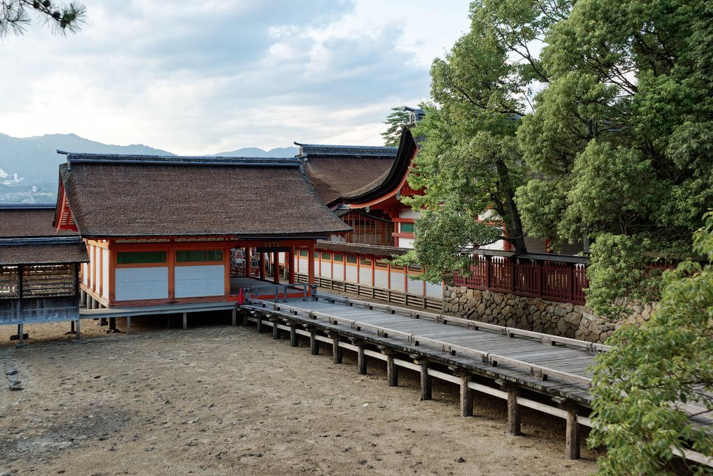 Red and white timber shrine buildings with dark roofs connect by raised corridors across a tidal shore beside trees and mountains.