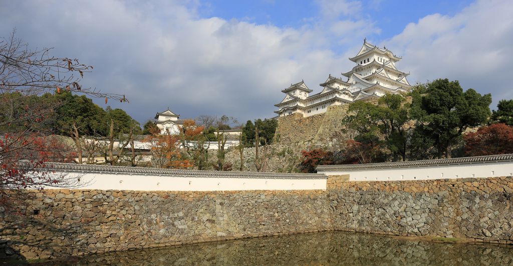 White multi-storey castle keeps rise above stone walls, a moat, trees and lower enclosures beneath broken cloud.