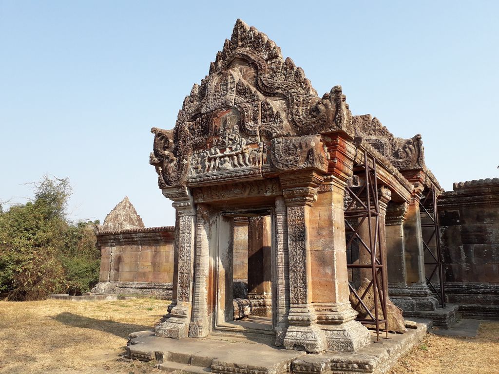 A richly carved weathered sandstone gateway stands in warm light with metal bracing and low temple walls behind.