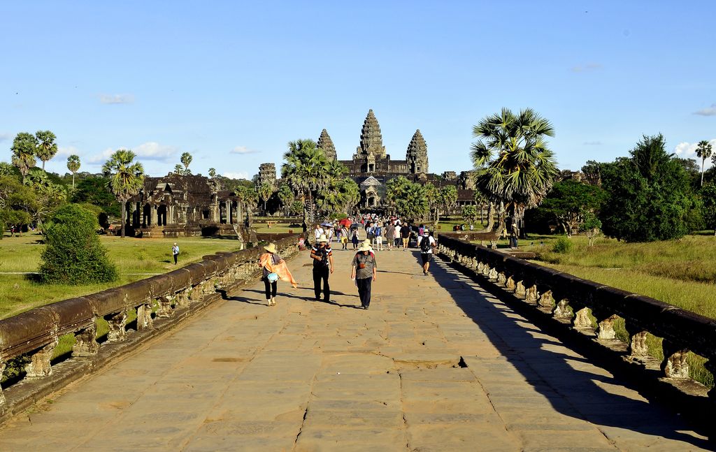 A broad paved causeway crowded with visitors leads between low stone balustrades toward Angkor Wat's towers.