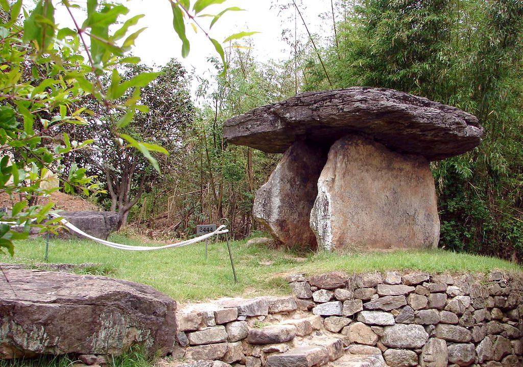 A massive flat stone cap rests on two upright stones in a wooded grass clearing beside a low retaining wall.