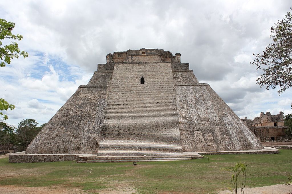 A steep central stair climbs a broad rounded stone pyramid under broken clouds, with low ruins and trees at its sides.