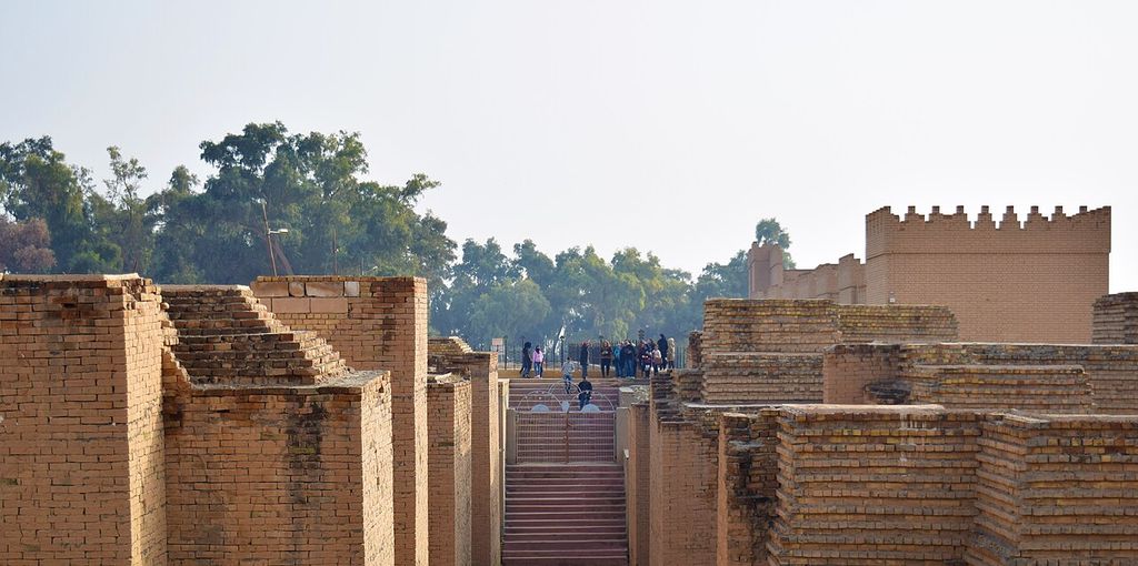 Layered tan brick walls flank a modern stair leading toward visitors and a high crenellated reconstructed wall among trees.