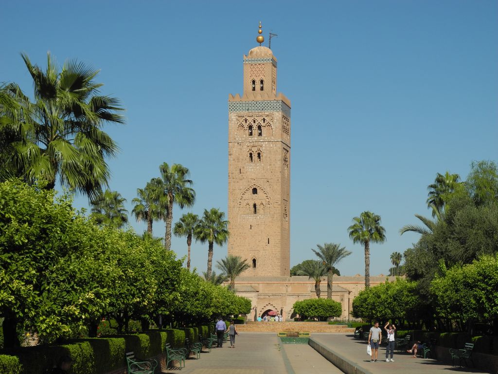 A tall square ochre minaret with geometric ornament rises behind palms, citrus trees, paths and visitors.